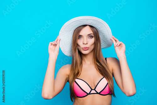 Portrait of lovely romantic girl with long hair holding two hands on white hat blowing sending air kiss with pout lips to the camera isolated over blue background