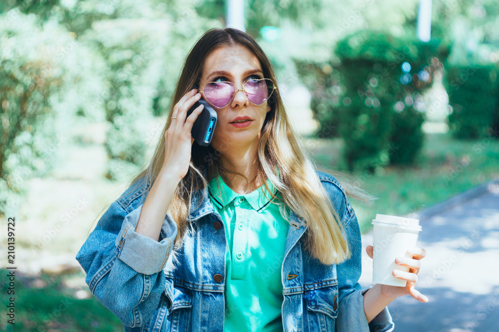 Attractive young girl is smiling and standing with a mug of coffee and a smartphone. Fashionable hipster model in trendy heart-shaped sunglasses. Concept of messaging and texting in the park Attractive young girl is smiling and standing with a mug of coffee and a smartphone. Fashionable hipster model in trendy heart-shaped sunglasses. Concept of messaging and texting in the park