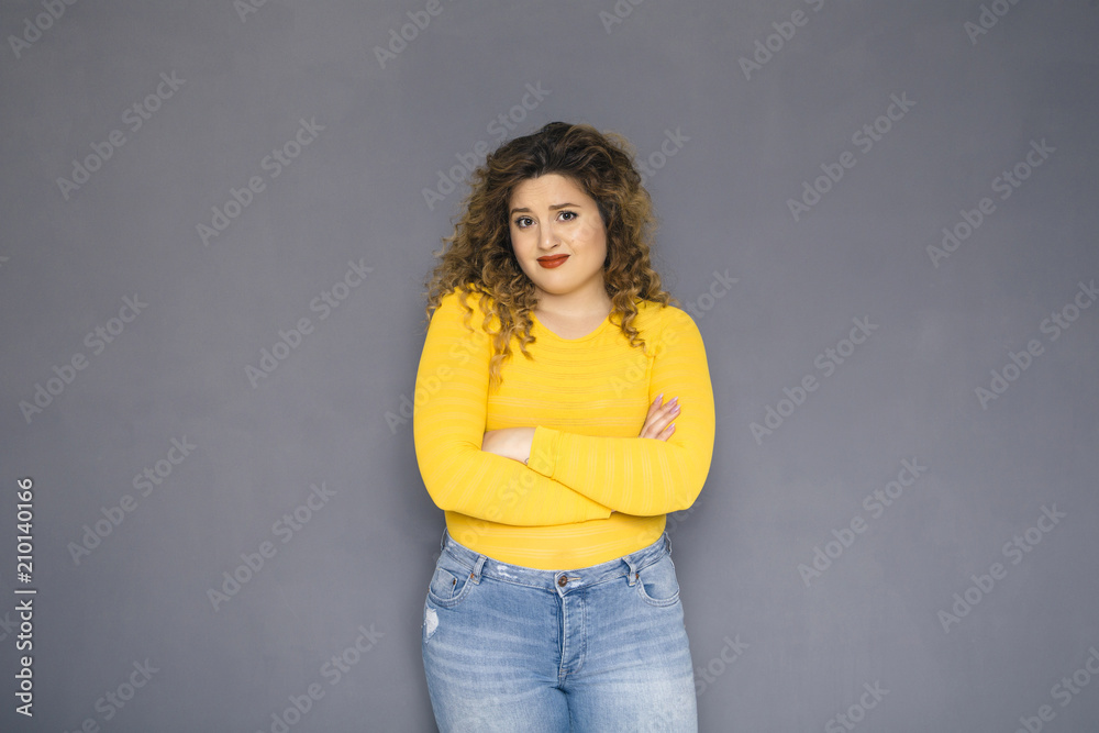 Cute brunette plus size woman with curly hair in yellow sweater and jeans standing on a neutral grey background. She doubt something, unhappy emotions on her face Cute brunette plus size woman with curly hair in yellow sweater and jeans standing on a neutral grey background. She doubt something, unhappy emotions on her face