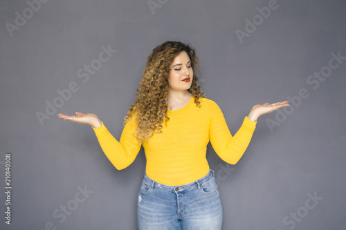 Cute brunette plus size woman with curly hair in yellow sweater and jeans standing on a neutral grey background. She is trying to decide what to choose. Holding hands and palms like plate for options