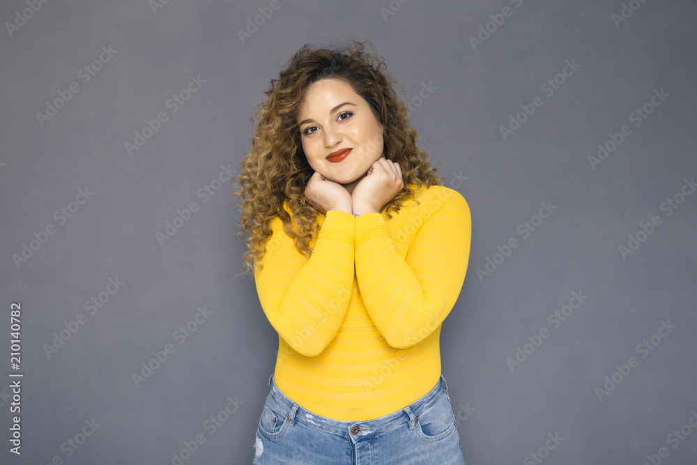 Cute brunette plus size woman with curly hair in yellow sweater and jeans standing on a neutral grey background. She was thinking, thoughtful emotion on her face. Copy space Cute brunette plus size woman with curly hair in yellow sweater and jeans standing on a neutral grey background. She was thinking, thoughtful emotion on her face. Copy space