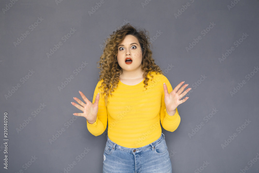 Cute brunette plus size woman with curly hair in yellow sweater and jeans standing on a neutral grey background. She amazed, shocked about some surprise