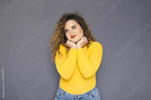 Cute brunette plus size woman with curly hair in yellow sweater and jeans standing on a neutral grey background. She was thinking, thoughtful emotion on her face. Copy space