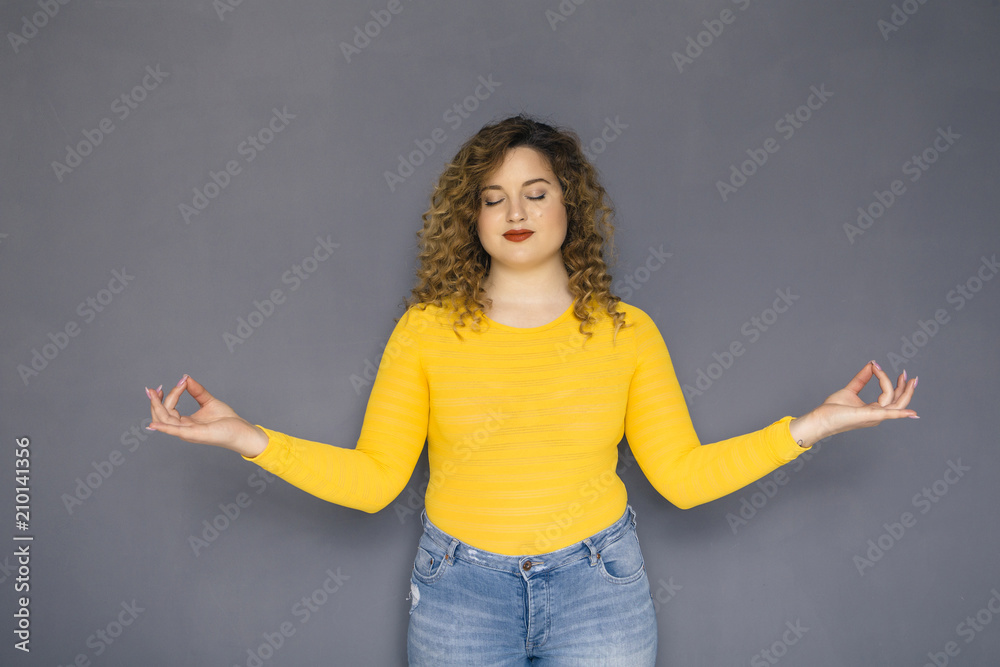 Cute brunette plus size woman with curly hair in yellow sweater and jeans standing on a neutral grey background. She makes a meditation gesture or sign with her hands Cute brunette plus size woman with curly hair in yellow sweater and jeans standing on a neutral grey background. She makes a meditation gesture or sign with her hands