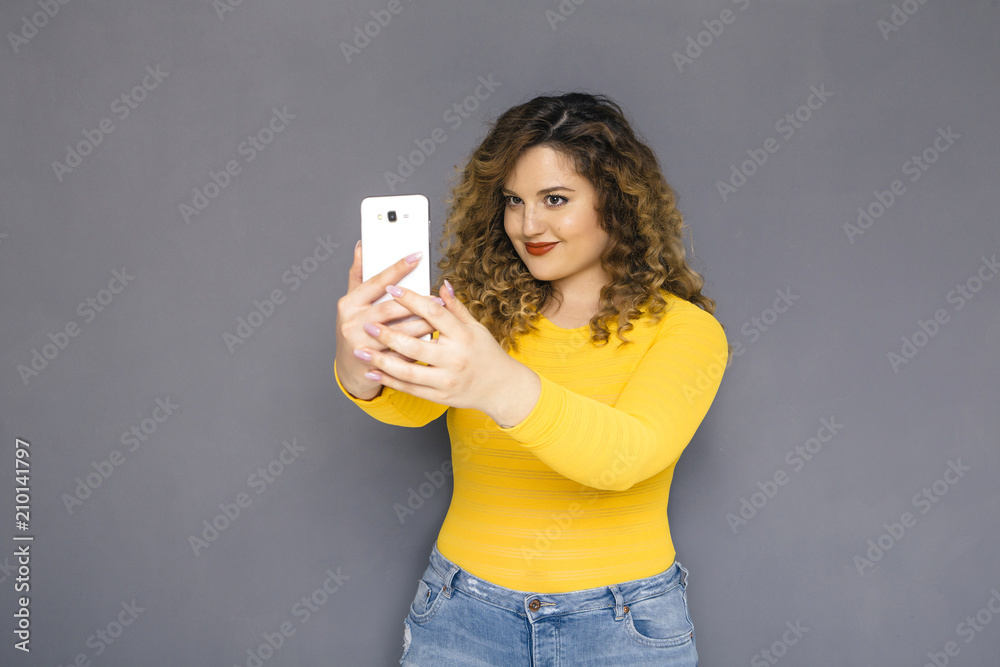 Cute brunette plus size woman with curly hair in yellow sweater and jeans standing on a neutral grey background. She makes selfie, photographing herself on her mobile phone. Copy space Cute brunette plus size woman with curly hair in yellow sweater and jeans standing on a neutral grey background. She makes selfie, photographing herself on her mobile phone. Copy space