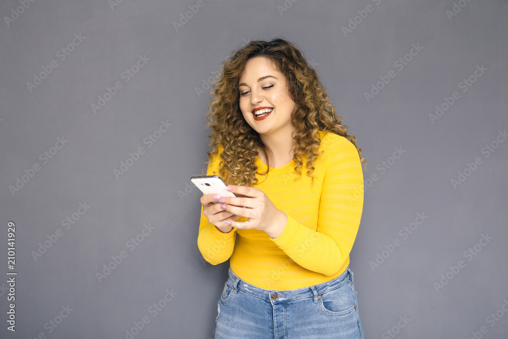 Cute brunette plus size woman with curly hair in yellow sweater and jeans standing on a neutral grey background. She chat on her mobile phone, typing an sms, amazed of what she read on it Cute brunette plus size woman with curly hair in yellow sweater and jeans standing on a neutral grey background. She chat on her mobile phone, typing an sms, amazed of what she read on it