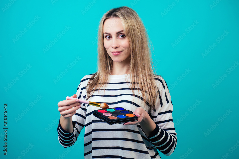 young and attractive blonde woman visagiste (make-up artist ) holds a brush make-up and a palette of eye shadows on blue background in a studio. concept of skin care and beauty cosmetics for face young and attractive blonde woman visagiste (make-up artist ) holds a brush make-up and a palette of eye shadows on blue background in a studio. concept of skin care and beauty cosmetics for face