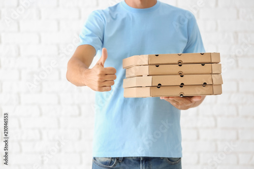 Young man with pizza boxes showing thumb-up gesture against white brick wall. Food delivery service