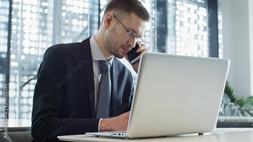 PAN shot of young handsome businessman in eyeglasses sitting at table in coworking space, writing notes and talking on mobile phone PAN shot of young handsome businessman in eyeglasses sitting at table in coworking space, writing notes and talking on mobile phone
