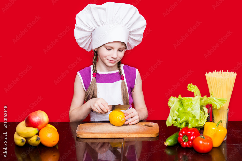 Happy little girl in chef uniform cuts fruit in kitchen. Kid chef. Cooking Process Concept Happy little girl in chef uniform cuts fruit in kitchen. Kid chef. Cooking Process Concept