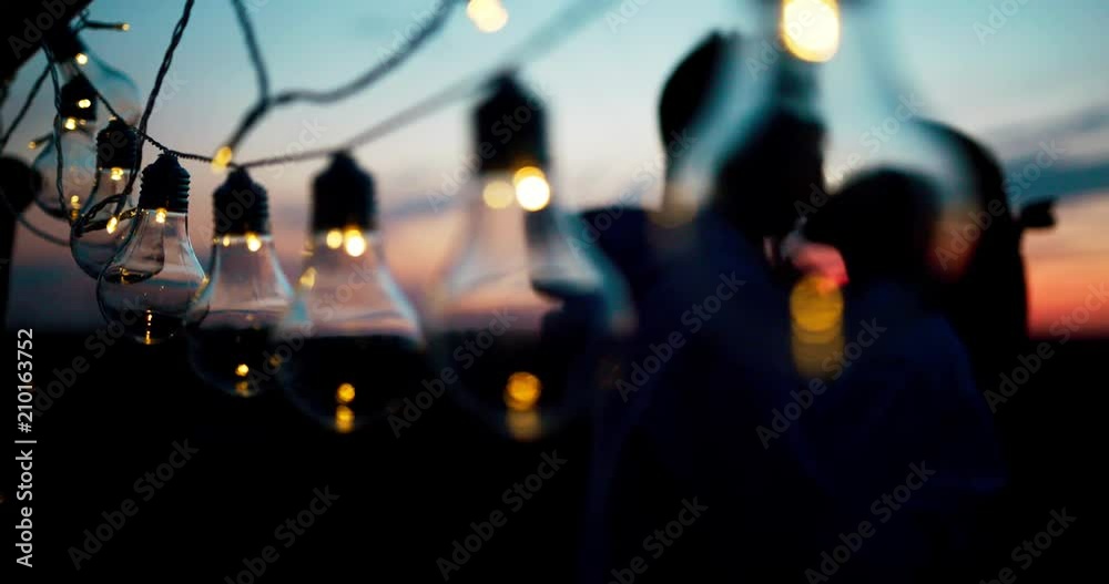 Romantic portrait of the hugging loving couple behind the string of lights during the sunset. 4k footage.
