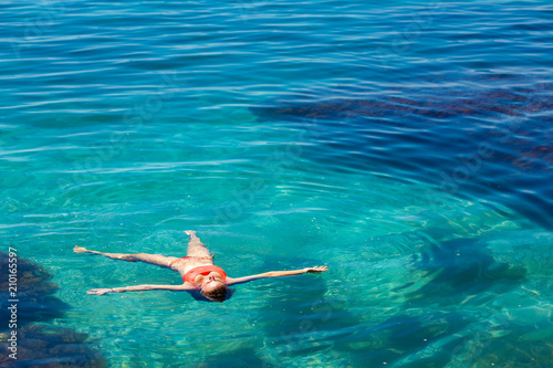 Vacation at sea. A young girl in a colored swimsuit lies an asterisk in a transparent ocean with turquoise water. Through the purity of the water  you can see the bottom with algae. Crimea. Black Sea.