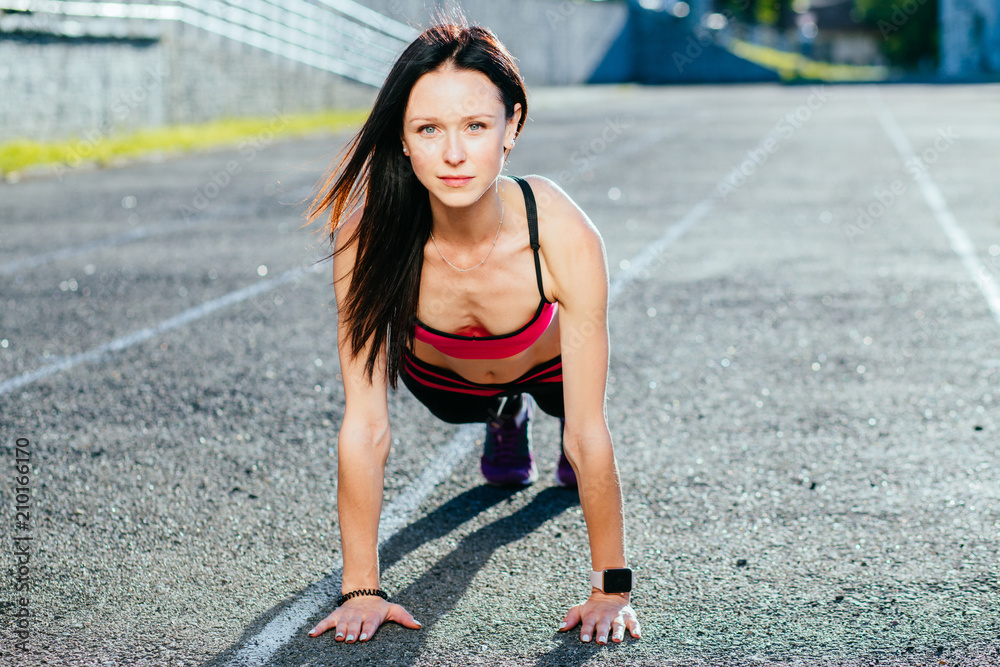 Happy gorgeous active brunette woman doing push up on stadium track in sunny summer day. Concentrated female doing physical drill concept.