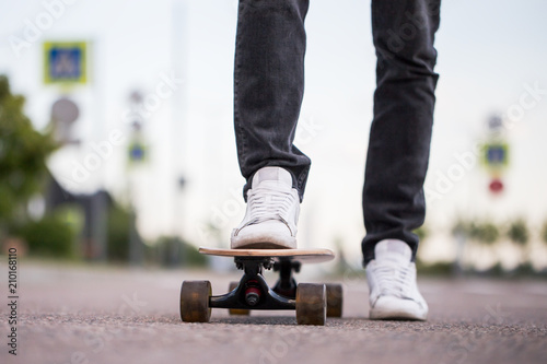 Closeup of man's leg in white sneakers ready to ride her wooden long board outdoors, front view, cropped image. Leisure, healthy lifestyle, extreme sports concept.Teenage male practicing long board
