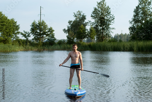 A man stands on a SAP Board with an oar and swims through a forest lake,
