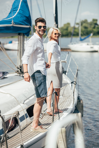 Beautiful happy young couple in sunglasses holding hands and smiling at camera on yacht