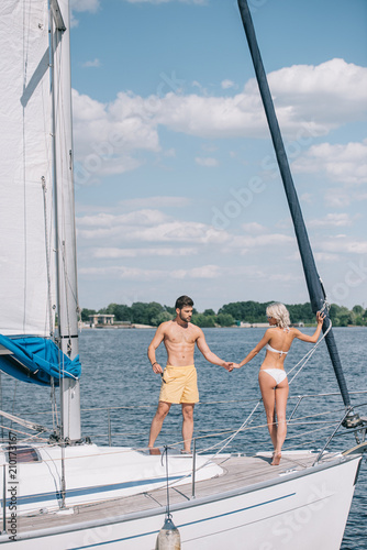 beautiful young couple in swimwear holding hands while standing together on yacht