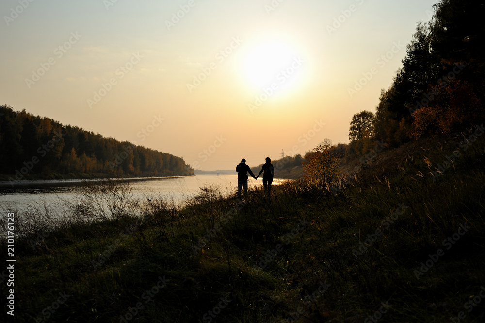 Young couple is walking along the river bank at sunset. Warm autumn evening. Silhouettes of a girl and a boy walking by the hand. Silhouettes of trees on the river bank and low setting sun in the sky.
