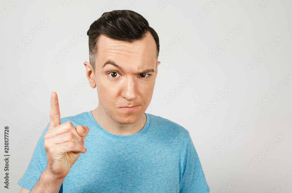 Young gloomy guy with finger up, dressed in a blue t-shirt on a light background. Close up portrait. Young gloomy guy with finger up, dressed in a blue t-shirt on a light background. Close up portrait.