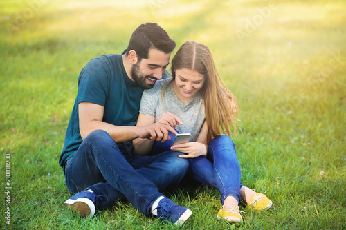 Happy young couple with mobile phone sitting on green grass in park © Pixel-Shot