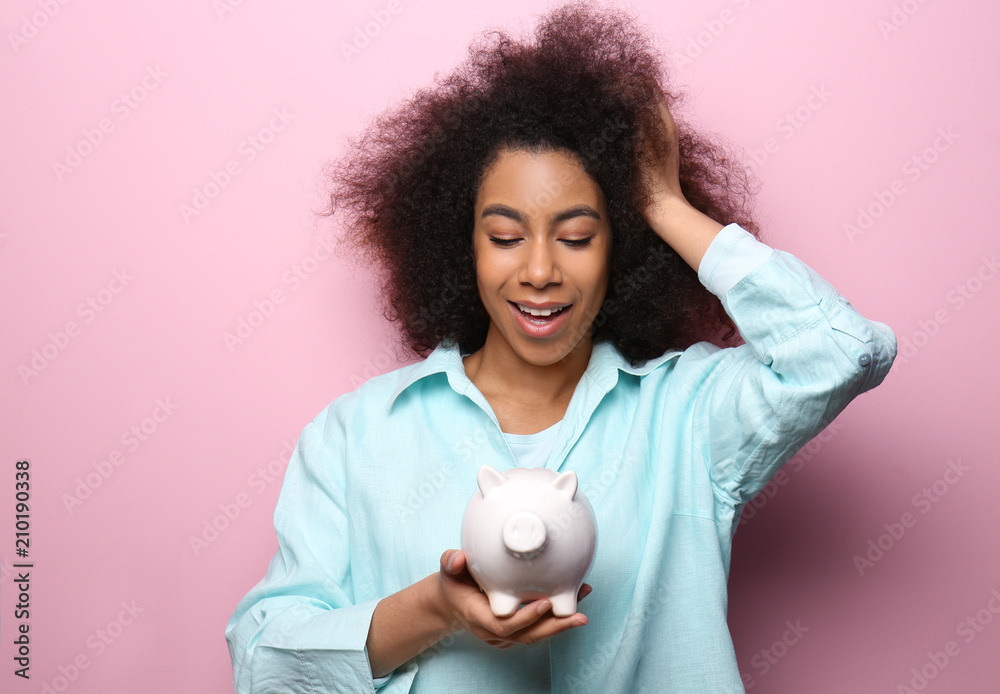 Young African-American woman with piggy bank on color background. Savings concept Young African-American woman with piggy bank on color background. Savings concept