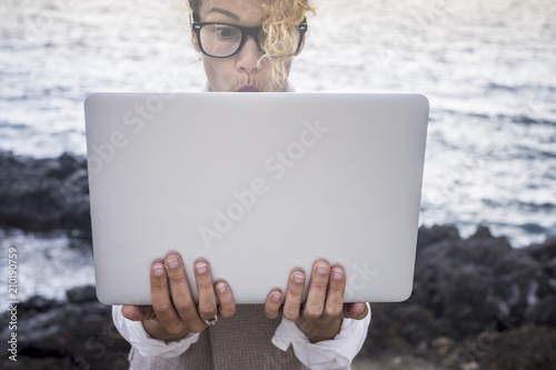 surprised face expression for beautiful caucasian young woman work on a laptop in alternative office outdoor on the coastline near the ocean waves. digital noman working outdoor concept photo