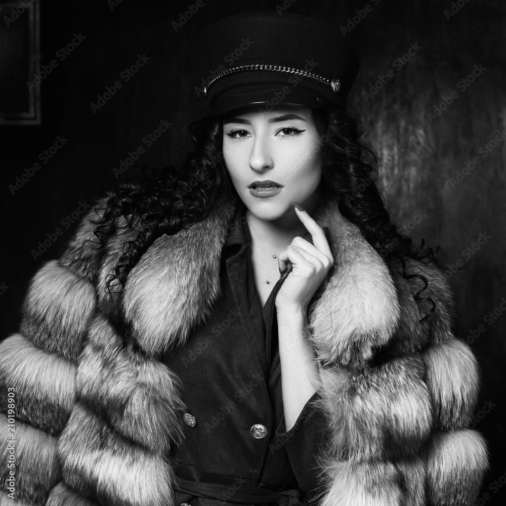 Stylish woman wearing green jacket dress and cap, fur coat and black jackboots posing in dark interior. Fashion style portrait. Brunette woman with curly hair. Stylish woman wearing green jacket dress and cap, fur coat and black jackboots posing in dark interior. Fashion style portrait. Brunette woman with curly hair.
