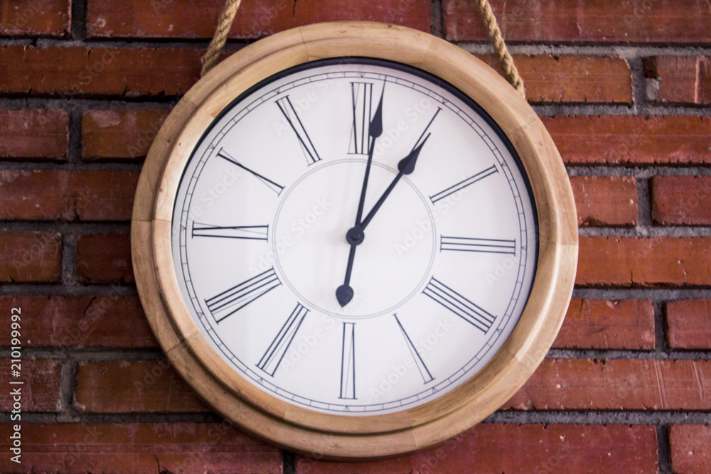 Close-up of a wooden classic and simple wall clock hanging in a red brick wall by a rope. It has roman numerals. Eye-level shot. Close-up of a wooden classic and simple wall clock hanging in a red brick wall by a rope. It has roman numerals. Eye-level shot.