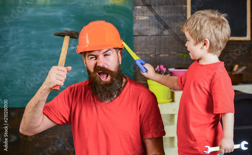 Fatherhood concept. Father, parent with beard in protective helmet teaching little son to use different tools in school workshop. Boy, child cheerful playing with toy saw, learning use tools with dad.