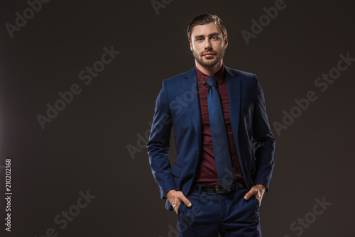 handsome businessman in stylish suit standing with hands in pockets and looking at camera isolated on black