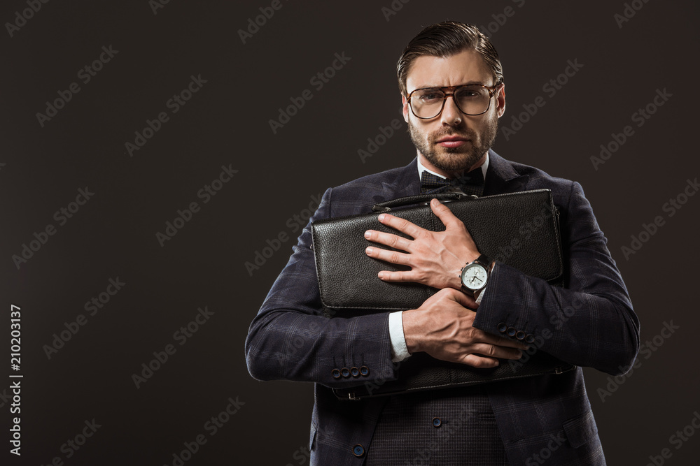 serious businessman in eyeglasses hugging briefcase and looking at camera isolated on black