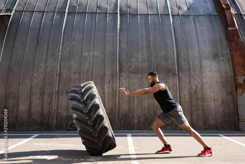 Fototapeta Naklejka Na Ścianę i Meble -  side view of athletic african american man exercising with tire on street