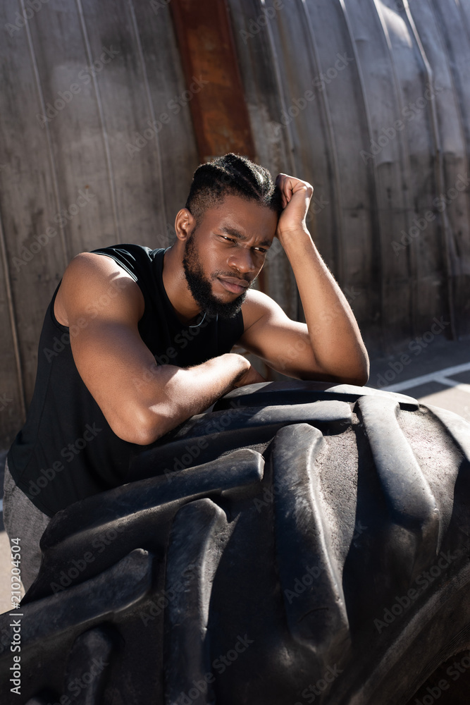 pensive african american sportsman leaning at tyre and looking away on street