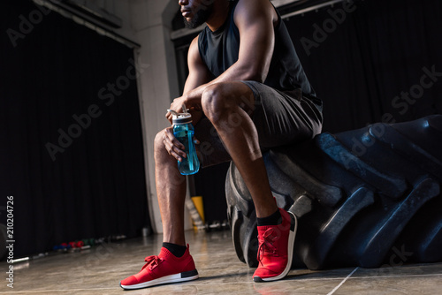 cropped shot of young african american sportsman sitting  tyre and holding bottle of water in gym