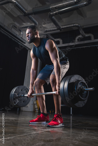 low angle view of muscular african american sportsman lifting barbell and looking away in gym
