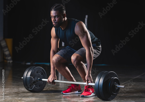 focused muscular african american sportsman lifting barbell and looking away in gym