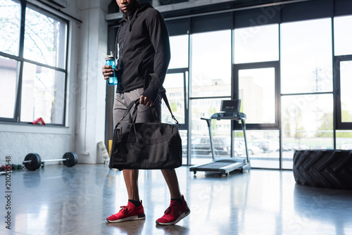 cropped shot of african american man in sportswear holding bag and bottle of water in gym photo