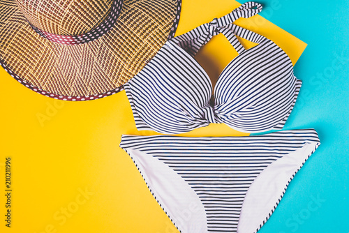 Top view of two pieces striped blue and white swimming suit and straw beach hat on blue and yellow pastel background. Concept of vacation.