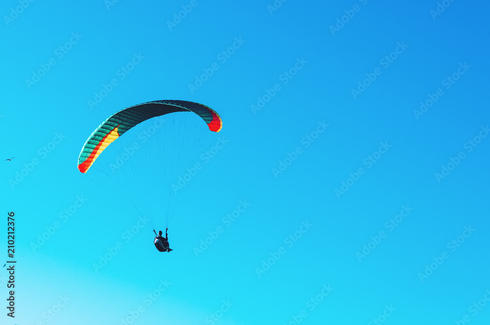 Paraglider flying on colorful parachute in blue clear sky at a bright sunny summer day. Active lifestyle, extreme sport. Adrenaline concept. Paraglider flying on colorful parachute in blue clear sky at a bright sunny summer day. Active lifestyle, extreme sport. Adrenaline concept.