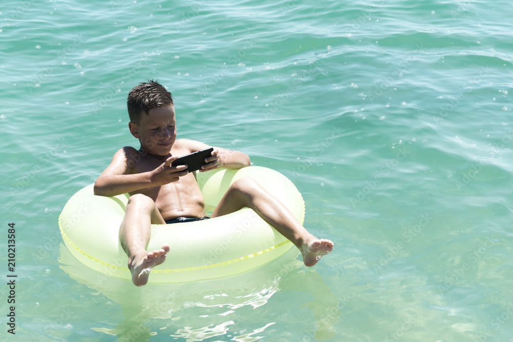 Boy lying on a rubber disc in the water and playing on the phone