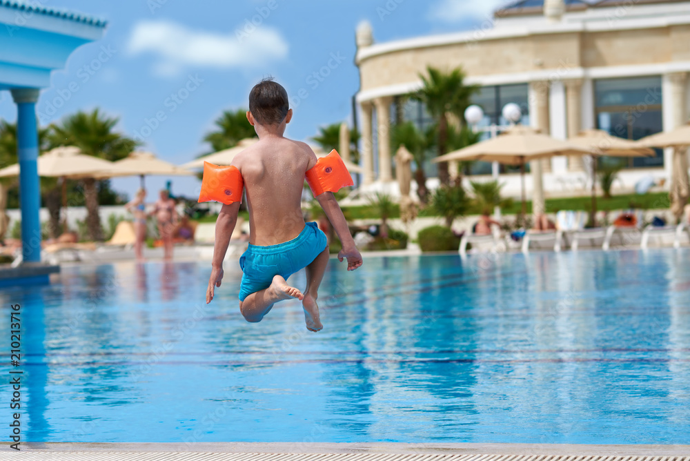European boy in floating sleeves having fun jumping into swimming pool at resort. Back view.