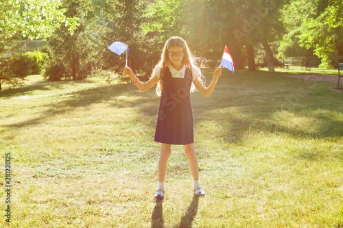 Cheerful charming little schoolgirl holding two flags in hands European Union and Netherlands. Background city park