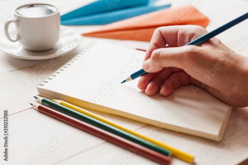 A hand with a color pencil is going to draw on a blank sheet of paper. Paper airplanes and a cup of coffee in the background. The concept of a dream of traveling, planning a vacation. Selective focus.