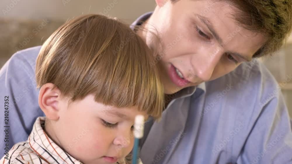 Young father and his little son painting on paper and holding tablet, dad helping and teaching his son how to write Young father and his little son painting on paper and holding tablet, dad helping and teaching his son how to write