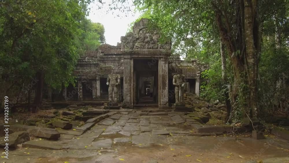 POV: Walking towards a large stone entrance to remnants of Buddhist temples protected by two decaying statues. Stunning view of a decaying religious building in Angkor Wat as people walk around halls POV: Walking towards a large stone entrance to remnants of Buddhist temples protected by two decaying statues. Stunning view of a decaying religious building in Angkor Wat as people walk around halls