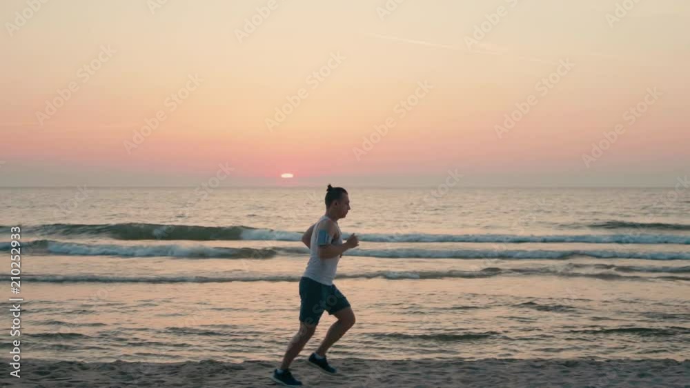 Sporty Hadsome Man is Jogging on Beach with Headphones, Running along Ocean Coast at Sunset or Sunrise in SlowMotion Sporty Hadsome Man is Jogging on Beach with Headphones, Running along Ocean Coast at Sunset or Sunrise in SlowMotion