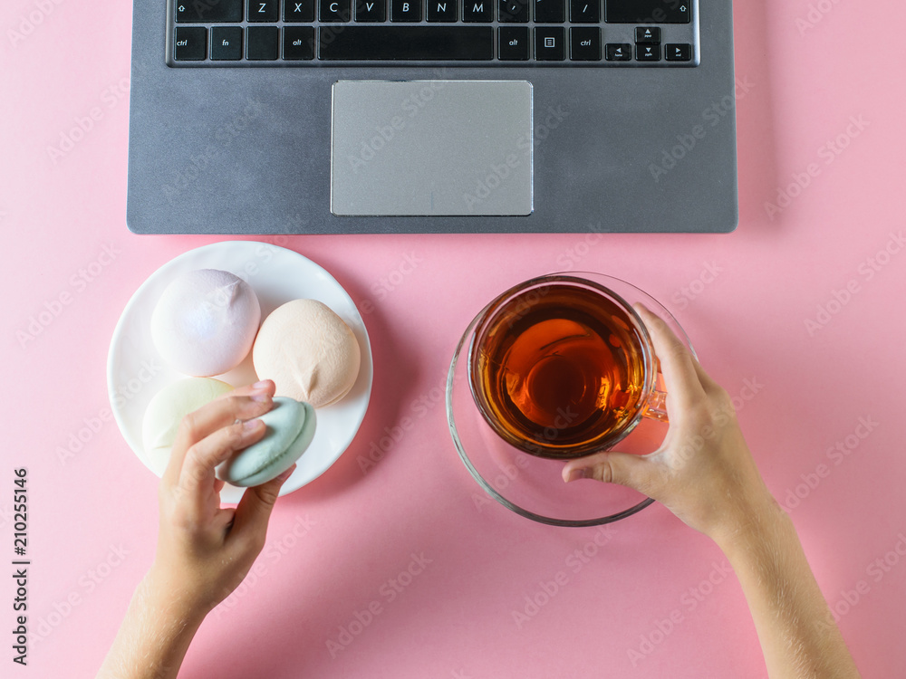 The girl eats colored marshmallows and drinks tea near the computer on a pink table. The view from the top.