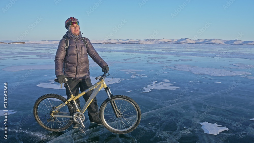 Man and his bicycle on ice. He stand and looks at the beautiful sunset. The cyclist is dressed in a gray down jacket, backpack and helmet. Ice of the frozen Lake Baikal. The tires on the bicycle are