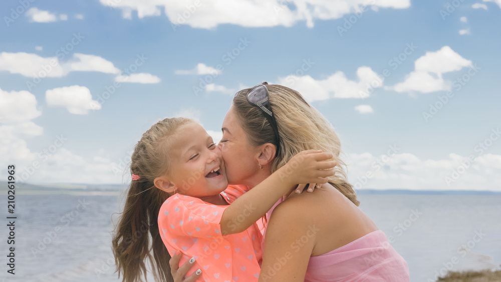 Little cute girl baby and young mother at beach. Mom sits on the shore, and her daughter is coming from behind. The kid hugs and kisses her mother. Mother and her child. Little cute girl baby and young mother at beach. Mom sits on the shore, and her daughter is coming from behind. The kid hugs and kisses her mother. Mother and her child.