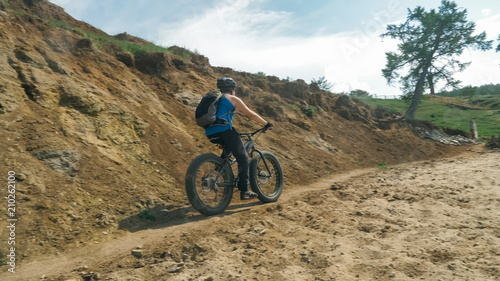 Fat bike also called fatbike or fat-tire bike in summer driving on the road. The guy rides by the hill on a sand clay path, behind him the shore by the sea.